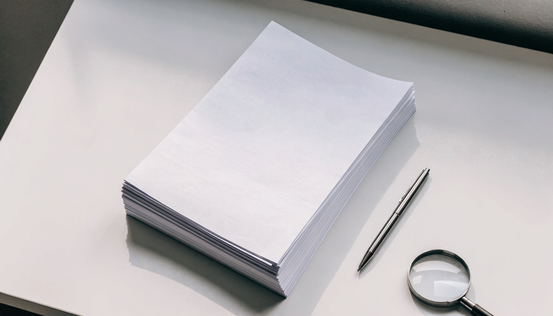 A neat stack of plain printed report sheets on a light grey desk beside a slim metal pen and a magnifying loupe
